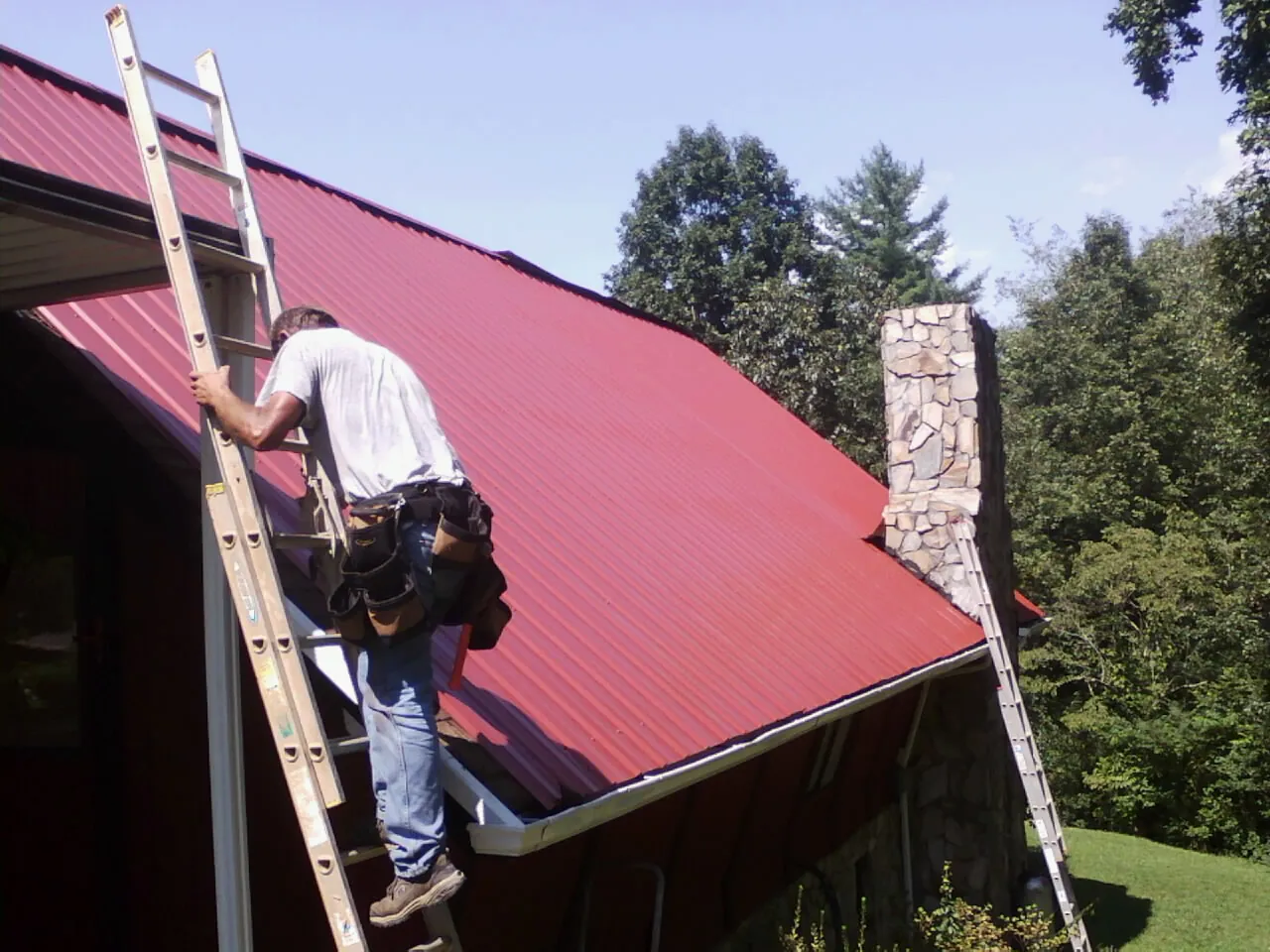 Roofer on a ladder beside a red metal roof and stone chimney — Soffit & Fascia Repair by Roofing Brothers in Harshaw